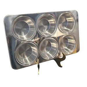 May include: A rectangular silver-colored metal muffin pan featuring six circular cups arranged in two rows of three. The pan has a smooth, reflective surface and is supported by a gold-toned metal display stand with curved legs.