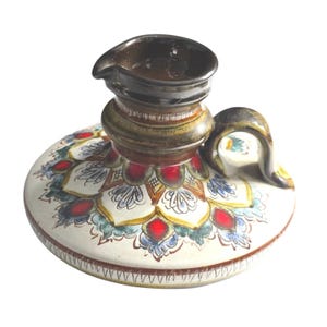 May include: A ceramic candle holder with a dark brown glazed top and handle. The base features a cream-colored surface decorated with a hand-painted floral mandala pattern in shades of red, blue, and gold, finished with a dark brown rim detail.