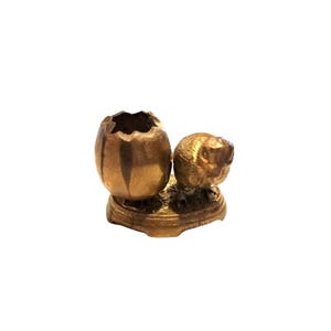 May include: A small, antique brass sculpture featuring a cracked egg and a small animal figure. The egg has a textured surface and a jagged opening. The animal figure is detailed and sits next to the egg on a round base.