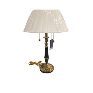 May include: A table lamp featuring a white pleated fabric shade, a slender black central column, and a brass-toned base with a leaf-like embossed pattern. The lamp includes two pull chains with small finials and a power cord with a plug.