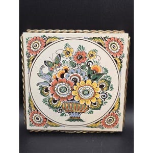 May include: Square ceramic tile with a floral design in a gold metal frame. The tile features a central bouquet of colorful flowers and a butterfly, surrounded by a decorative border. The frame has a twisted rope design.