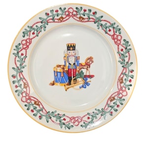 May include: A round white ceramic dinner plate with a gold rim. The center features a colorful nutcracker, a blue drum, a toy horse, and a gold trumpet. The border is decorated with a repeating pattern of green holly leaves and red berries.