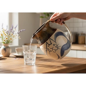 May include: A ceramic pitcher with a cat design is pouring water into a glass. The pitcher has a brown and beige color scheme with blue accents. The glass is clear, and the water is sparkling.
