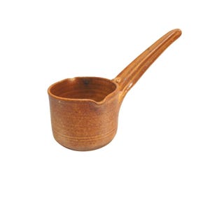May include: A handmade, brown ceramic ladle with a long, slender handle. The ladle has a small, round bowl with a pouring spout. The ceramic has a textured surface and a warm, earthy tone, suggesting a rustic or farmhouse style.