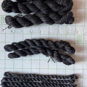 May include: Close-up of several skeins of dark gray yarn, arranged on a grid. The yarn appears to be a twisted, textured fiber, suitable for knitting or crocheting projects. The image shows different yarn weights and textures.