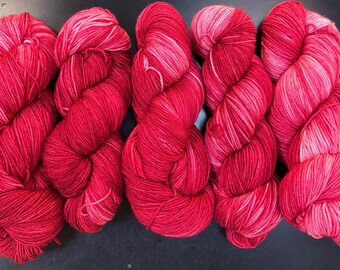 May include: Five skeins of vibrant red yarn with subtle pink and white highlights. The yarn is tightly wound, showcasing a textured appearance. Ideal for knitting or crocheting projects, the yarn is arranged on a dark surface.