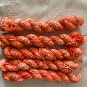 May include: Four skeins of hand-dyed yarn in peach and cream hues. The yarn is twisted into a rope-like form, with the colours blending. The yarn is likely wool or a similar fibre, suitable for knitting or crochet projects. Each skein is approximately 100g.