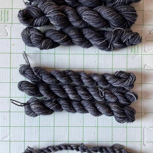 May include: Several skeins of dark gray yarn with subtle white and light gray marbling. The yarn is tightly twisted, showing a textured surface. The image is taken from above, showing the yarn against a grid background.
