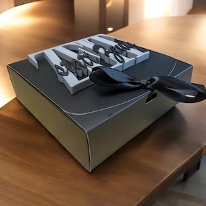 Könnte beinhalten: Eine schwarze Geschenkbox mit einem grau-schwarzen Dekorationselement. Das Element schreibt "ZARA Engaged". Die Box ist mit einem schwarzen Band gebunden. Auf einer Holzoberfläche.