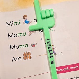 Op de afbeelding: Een felgroene, handvormige leeswijzer ligt op een pagina van een boek. De aanwijzer heeft de tekst "LESEBÄR M" erop gedrukt. De pagina van het boek heeft de woorden "Mimi", "Mama" en "Am".