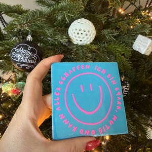 Könnte beinhalten: Eine quadratische, hellblaue Kachel mit einem pinkfarbenen Smiley und den Worten "ALLES SCHAFFEN ICH BIN STARK, MUTIG UND KANN" in einem Kreis. Die Kachel wird vor einem Weihnachtsbaum mit Ornamenten gehalten.
