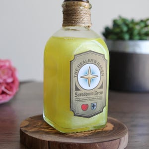 Puede incluir: Una botella de vidrio transparente llena de un líquido amarillo, etiquetada como "The Healer's Haven Saradomin Brew". La botella tiene un tapón de corcho envuelto con cuerda y se encuentra sobre un posavasos de madera. La etiqueta incluye una rosa de los vientos y un símbolo de corazón.