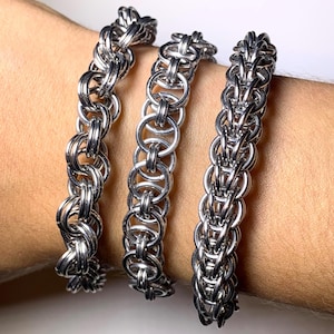 Pode incluir: Três pulseiras de cota de malha prateadas. Cada pulseira apresenta um design de tecido de cota de malha diferente. As pulseiras são exibidas em um pulso, destacando o intrincado trabalho em metal e as diversas texturas das joias.