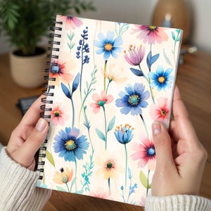 Könnte beinhalten: Ein spiralgebundenes Notizbuch mit floralem Design. Das Cover zeigt ein Aquarellmuster aus bunten Blumen in Blau-, Rosa- und Gelbtönen auf cremefarbenem Hintergrund. Das Notizbuch ist geöffnet und zeigt den lebendigen Blumendruck.