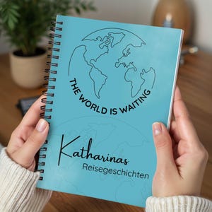 Könnte beinhalten: Ein spiralgebundenes Notizbuch mit türkisblauem Einband, das einen schwarzen Umriss der Weltkarte und den Text "THE WORLD IS WAITING" zeigt. Darunter stehen der Name "Katharinas" und die Worte "Reisegeschichten" in Schwarz.
