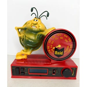 May include: A red and black novelty AM/FM radio featuring a yellow and green cartoon bug character leaning against a circular red dial. The radio includes a digital display, a volume knob, and the Raid logo printed on the center of the dial.