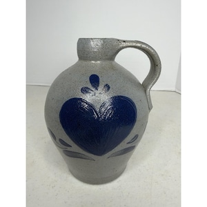 Puede incluir: Una jarra de gres gris con textura moteada y una sola asa. La parte frontal presenta un diseño de corazón azul cobalto prominente con detalles decorativos en forma de hoja por encima y por debajo, todo en un esmalte azul brillante.