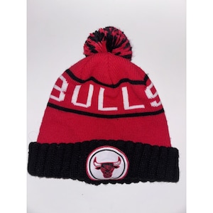 May include: A red and black knit beanie featuring a pom-pom on top. The word BULLS is knitted in white letters across the center. A circular patch with a red and black bull logo is centered on the thick black ribbed cuff at the bottom edge.