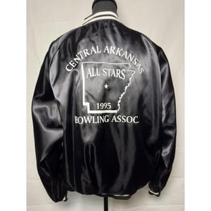 May include: A black satin bomber jacket featuring white striped ribbed trim at the collar. The back displays a white graphic of the state of Arkansas with the text CENTRAL ARKANSAS ALL STARS 1995 BOWLING ASSOC. printed in white lettering.