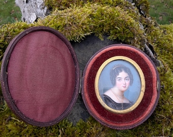 May include: An antique miniature portrait of a woman in a round, hinged case. The portrait features a woman with dark hair and a black dress, set against a blue background. The case is lined with burgundy velvet and has a dark brown exterior.