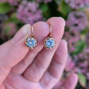 Peut inclure: Une paire de boucles d'oreilles pendantes en or avec une pierre précieuse ronde, claire et facettée, sertie dans une monture métallique décorative festonnée. Chaque boucle est suspendue à un crochet en or simple et incurvé.