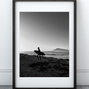 Puede incluir: Impresión enmarcada en blanco y negro de un surfista caminando por una playa de arena, llevando una tabla de surf. La silueta del surfista se recorta contra el océano y una cordillera distante. La imagen está enmarcada en un marco de madera oscura.
