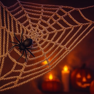 May include: A cream-colored, crocheted spiderweb with a black plastic spider attached. The Halloween decoration is set against a warm, orange background with lit candles and carved pumpkins.