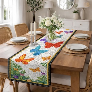 Pode incluir: Caminho de mesa acolchoado branco com um design de borboletas. O caminho de mesa apresenta borboletas em tons de laranja, vermelho, azul e roxo. As bordas são pretas com uma borda dourada. O caminho de mesa é colocado sobre uma mesa de madeira.