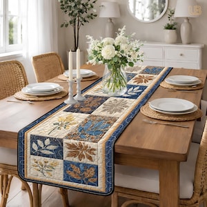 May include: A quilted table runner with a patchwork design featuring leaf patterns in shades of blue, brown, and cream. The runner is placed on a wooden dining table set with plates, candles, and a floral centerpiece.