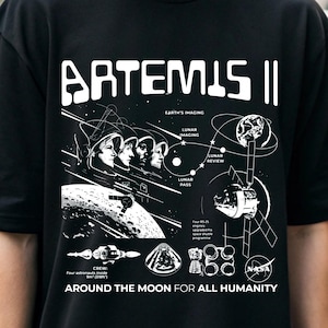 Puede incluir: Camiseta negra con un diseño gráfico blanco. El diseño incluye el texto "ARTEMIS II", ilustraciones de astronautas, la luna y diagramas espaciales, además del logotipo de la NASA. El texto "AROUND THE MOON FOR ALL HUMANITY" también está impreso.