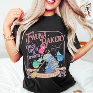May include: Black t-shirt featuring a whimsical design for "Fauna Bakery." The graphic includes the text "Once Upon A Dream" and "Baked with Love & Magic," along with illustrations of fairies and a cake. The bakery's established date, "Since 1959," is also displayed.