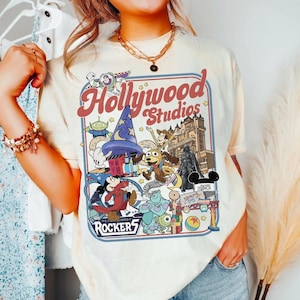 Może przedstawiać: Kremowa koszulka z kolorową grafiką Disney's Hollywood Studios. Projekt zawiera napis "Hollywood Studios" oraz różne postacie z kreskówek, takie jak Myszka Miki, Buzz Astral i Sully.