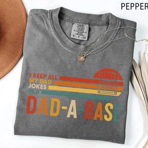 May include: Pepper-colored t-shirt with the text "I KEEP ALL MY DAD JOKES IN A DAD-A-BASE" in a retro design. The shirt has a crew neck and short sleeves. A silver necklace with a pendant is on the shirt.