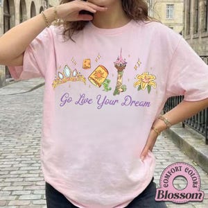 May include: A light pink t-shirt with a graphic featuring a gold tiara, a lantern, a tower, a flower, and the text "Go Live Your Dream" in purple. The shirt also has a Comfort Colors logo.