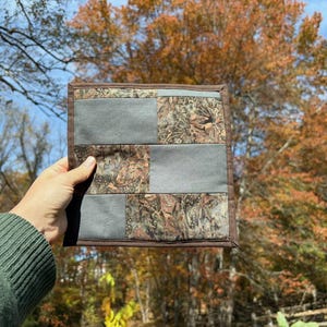 May include: A square, quilted fabric item with a camouflage pattern and gray rectangles. The item is held in a hand, with a brown border. The background features trees with fall foliage and a blue sky.