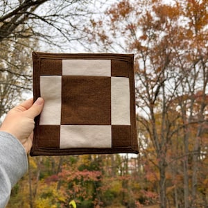 May include: A square, brown and cream-colored checkerboard-patterned hot pad. The hot pad is made of a soft material and has a stitched border. The background is a blurred view of trees with fall foliage.