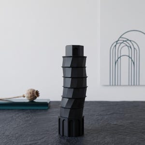 May include: A matte black sculptural object shaped like a leaning tower, featuring a series of stacked, slightly offset hexagonal segments. The base has vertical arched cutouts, and the top segment includes a small arched opening on its side.