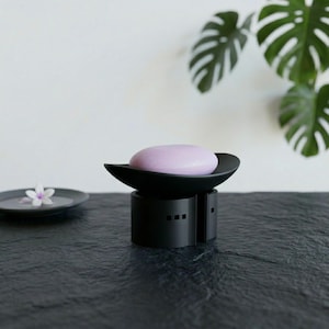 May include: A black soap dish with a lavender bar of soap. The dish has a unique design with a curved top and a cylindrical base. A small gray dish with a flower is in the background. The soap dish is on a dark surface.