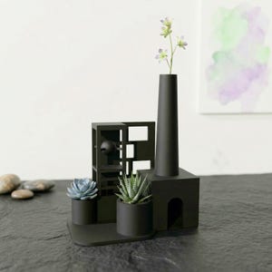May include: A modern black decorative sculpture with a tall vase holding flowers, a geometric structure, and two small potted succulents. The sculpture sits on a dark surface, with a watercolor painting in the background.
