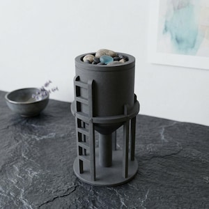 May include: A dark gray cylindrical container shaped like a water tower, featuring a vertical ladder on the side and a conical base. The top opening is filled with a collection of smooth, rounded river stones and a single translucent blue glass pebble.