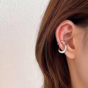 May include: A silver ear cuff featuring a curved design that wraps around the ear. The cuff has a smooth, polished finish and is worn on the upper ear. The jewelry is a modern accessory.
