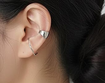 Puede incluir: Un brazalete de oreja plateado y un pequeño pendiente curvo se llevan en la oreja. El brazalete de oreja es un diseño simple y curvo que se envuelve alrededor de la parte superior de la oreja. El pendiente es una línea fina y ondulada.