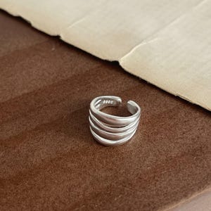 May include: A silver-colored ring with a layered design. The ring has a smooth, polished finish and features the text "S925" on the band. The ring is set against a brown background.