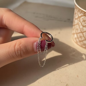 Könnte beinhalten: Ein silberner Ohrklemmen-Schmuck mit drei zarten Kettenanhängern. Der Ohrklemmen-Schmuck hat ein schlichtes, gebogenes Design, das sich um das Ohr legt. Die Ketten hängen herab und verleihen Bewegung und einen Hauch von Eleganz. Der Schmuck wird vor einem weichen, neutralen Hintergrund gezeigt.