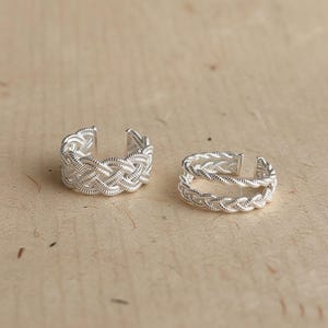 May include: Two silver-colored toe rings with a woven design. The rings are open at the top, allowing for an adjustable fit. The rings are displayed on a light brown surface.