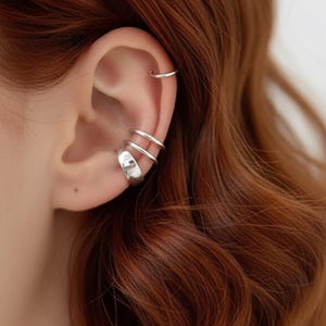 Könnte beinhalten: Nahaufnahme eines Ohrs, das mit silbernen Ohrklemmen und einem kleinen Creolen-Ohrring geschmückt ist. Die Ohrklemmen umfassen ein breites Band und zwei dünnere Bänder, wodurch ein geschichteter Effekt entsteht. Der Schmuck ist vor dem Hintergrund von rotem Haar platziert.