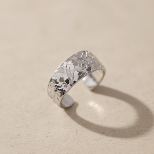 May include: A silver ring with a hammered texture. The open-ended ring features a wide band with an irregular pattern. The ring is displayed against a neutral background.