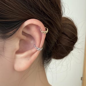 Puede incluir: Primer plano de una oreja adornada con dos puños de oreja de eslabones de cadena. Uno es dorado y está colocado en la parte superior de la oreja, mientras que el otro es plateado y está colocado más abajo. Fondo borroso.