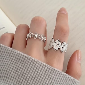 May include: Two silver rings with intricate lace-like designs. One ring features a floral pattern, while the other has a wider, more ornate lace design. The rings are displayed on a person's fingers, showcasing their delicate details.