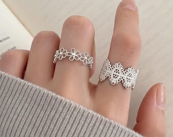 May include: Two silver rings with intricate lace-like designs. One ring features a floral pattern, while the other has a wider, more ornate lace design. The rings are displayed on a person's fingers, showcasing their delicate details.